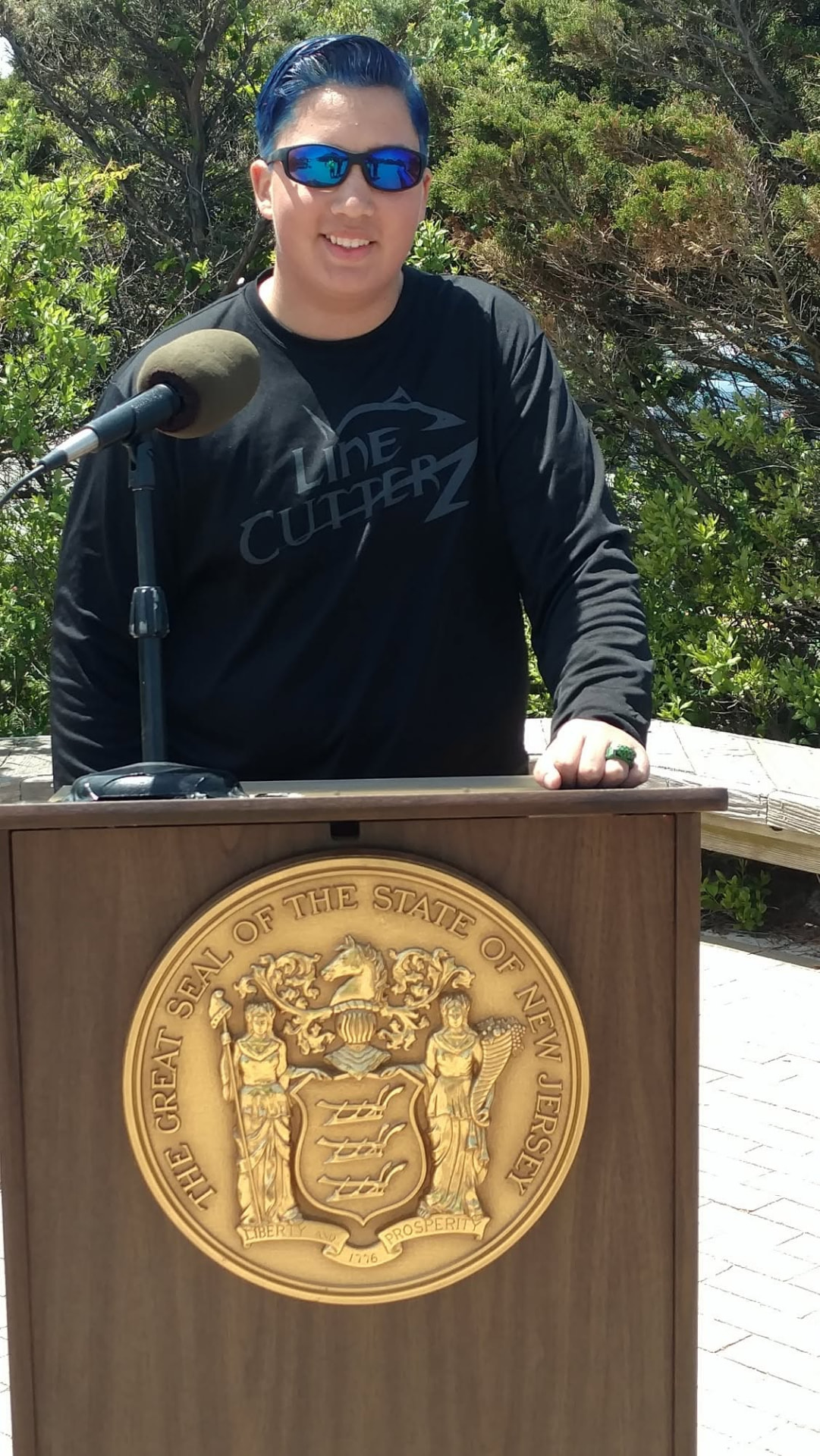 AJ-Max speaking at a podium bearing the Great Seal of the State of New Jersey
